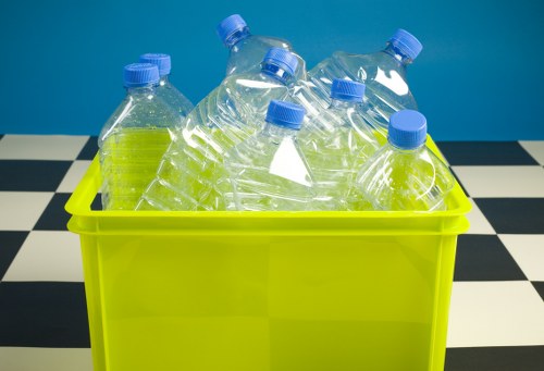 A bright yellow plastic container holding several empty clear plastic water bottles with blue caps, placed on a black-and-white checkered surface against a blue background. The bottles are arranged with some leaning against each other, and their transparent surfaces reveal the bottle shapes and ridged necks. The setting appears to be indoors, possibly in a commercial waste collection context. This image exemplifies waste items suitable for removal or recycling services provided by Commercial Waste Raynes Park, supporting proper disposal of plastic bottles in the local area near Raynes Park and the postcode region. The visual focus is on the packaging and environmental context for rubbish removal relevant to domestic or commercial waste management within the London Borough of Merton.