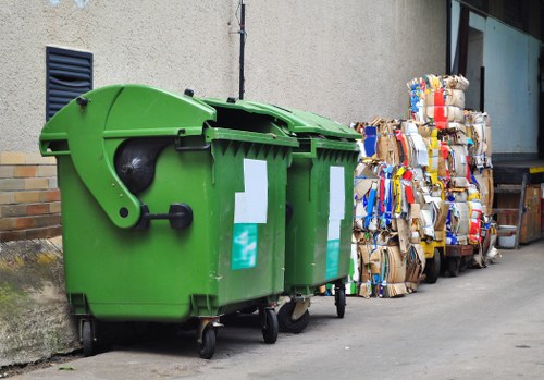In the foreground, there are two large green waste bins with black lids, positioned on a paved street near a concrete wall. The bins are made of durable plastic with textured surfaces, and they feature white recycling labels attached to their fronts. To the right of the bins, several tightly packed bundles of flattened cardboard boxes are stacked, showcasing various colors and patterns from the packaging, and are secured with tape or string. The arrangement suggests a commercial waste collection point, possibly in Raynes Park, with the bins and cardboard bundles ready for collection or disposal. The background includes a beige building facade with small, dark ventilation grilles, and a side window partially visible. The scene is evenly lit, indicating daytime with no visible shadows, and the overall environment appears clean and orderly, reflecting a routine rubbish removal process managed by Commercial Waste Raynes Park, a professional waste management service specializing in rubbish collection in the local area.