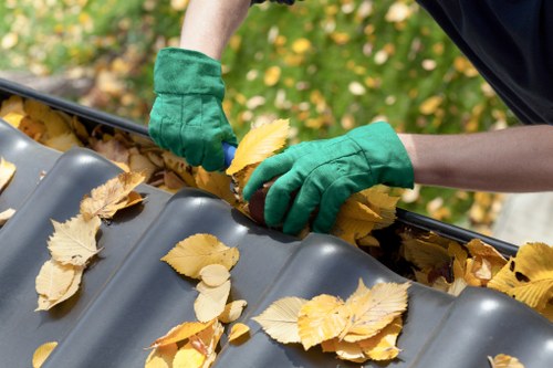 A person wearing green work gloves is seen collecting fallen yellow and brown leaves from a dark grey metal gutter installed on a building or roof structure during autumn, with a background of blurred foliage and greenery. The gutter has a smooth, slightly reflective surface and is partially filled with leaves, indicating ongoing maintenance or cleaning work. The person’s arms and hands are visible, demonstrating careful removal of debris. This scene is situated outdoors, possibly in a residential or commercial area in Raynes Park, as part of routine rubbish or waste management tasks by Commercial Waste Raynes Park, ensuring the proper clearance of garden debris to maintain property cleanliness.