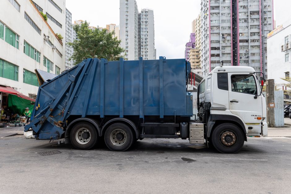 A large commercial waste collection truck is parked on an urban street, featuring a white cab and a blue, enclosed waste container at the rear. The container is made of metal with a smooth, slightly r