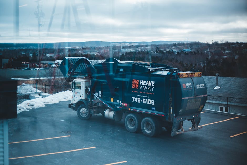 A large commercial waste collection truck is parked on an urban street, featuring a white cab and a blue, enclosed waste container at the rear. The container is made of metal with a smooth, slightly r